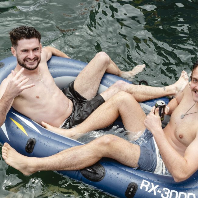 Two men lounging in an inflatable raft on a body of water. One is shirtless, making a hand gesture and smiling, while the other holds a drink. Both appear relaxed and are enjoying their time on the water.