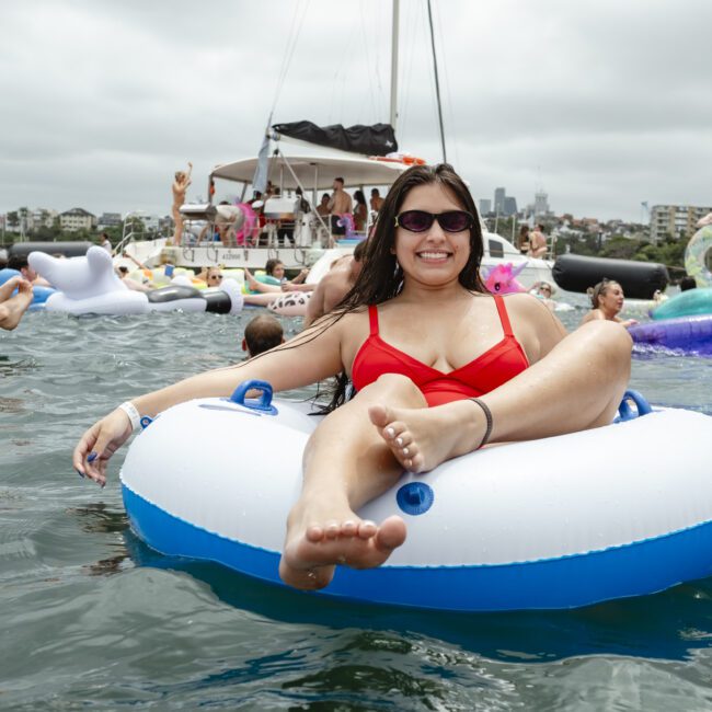 A person in a red swimsuit relaxes on an inflatable tube in a body of water. Other people on inflatables and boats are in the background, enjoying a festive, sunny atmosphere.