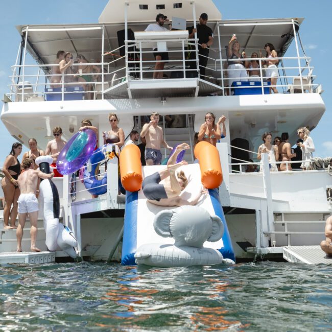 A group of people enjoy a sunny day on a boat, with some using inflatable slides and floats to enter the water. The scene is lively, with people watching, swimming, and relaxing on the multi-level vessel.