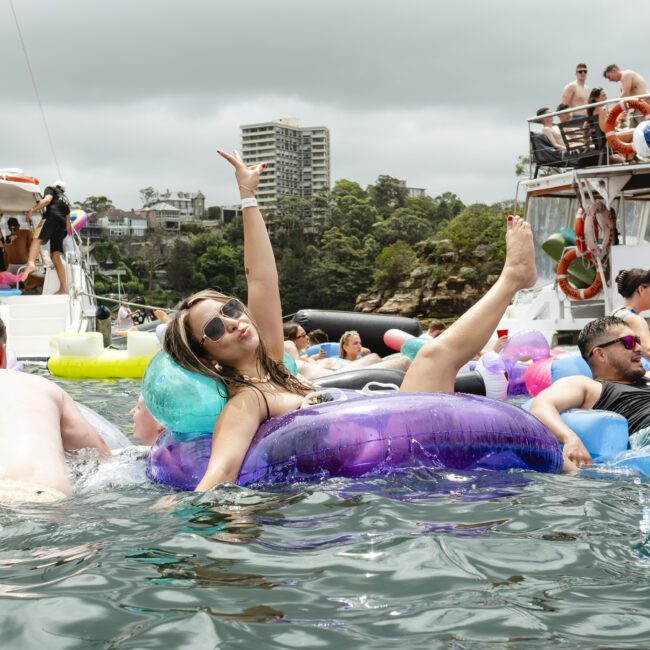 A group of people enjoying a boat party, lounging on colorful inflatable floats in the water. A woman wearing sunglasses strikes a playful pose with one arm and leg up. Boats and greenery are visible in the background under a cloudy sky.