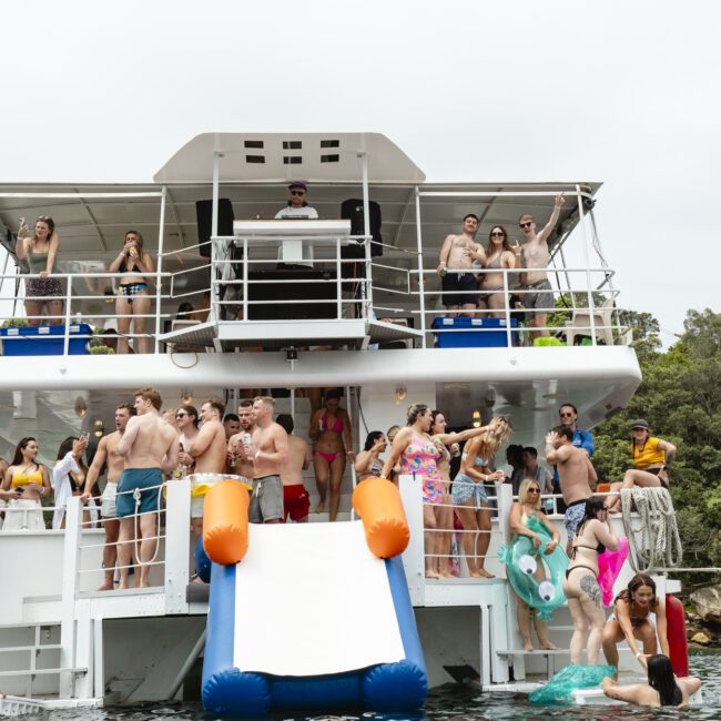 A group of people enjoy a boat party, with some standing on decks, others using an inflatable water slide, and several swimming. A person is captured mid-dive. A seagull flies overhead, and trees are visible in the background.