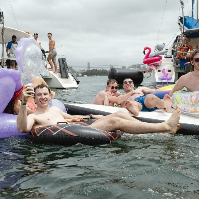 A group of people enjoying a day on the water, lounging on inflatable rafts. They are smiling and holding drinks, surrounded by boats. The atmosphere is relaxed and festive. Overcast skies set the backdrop.