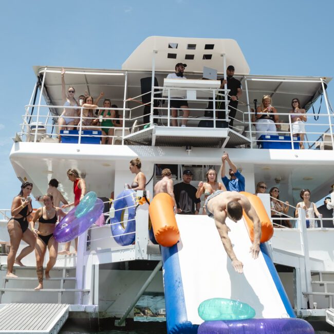 A group of people enjoying a boat party on a sunny day. Some are on the deck, while others are using a slide to enter the water. Inflatable tubes float nearby. The atmosphere is lively and festive.