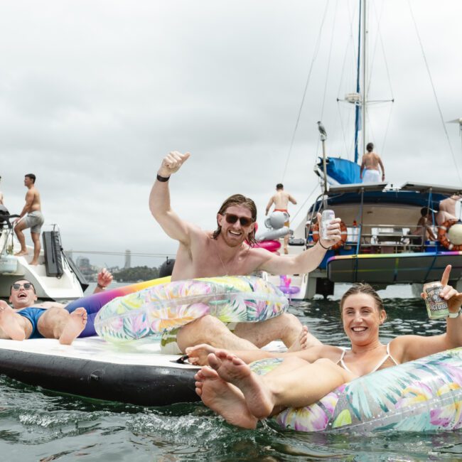 A group of people enjoying a day on inflatable tubes in the water, with sailboats in the background. They are smiling and holding drinks, creating a festive and relaxed atmosphere on a cloudy day.