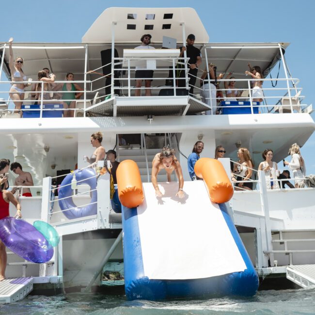 People are enjoying a party on a boat. A person is sliding down an inflatable slide into the water. Others are on the deck, some with drinks, under a clear blue sky.