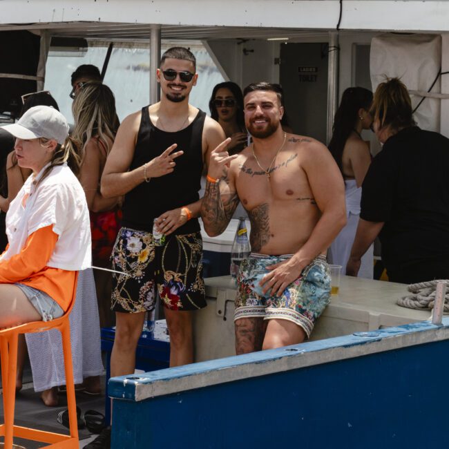 A group of people enjoying a boat trip. Two men in beachwear are posing and smiling for the camera. Others are sitting and relaxing in the background. The boat has a small seating area and some equipment visible. The atmosphere is casual and lively.