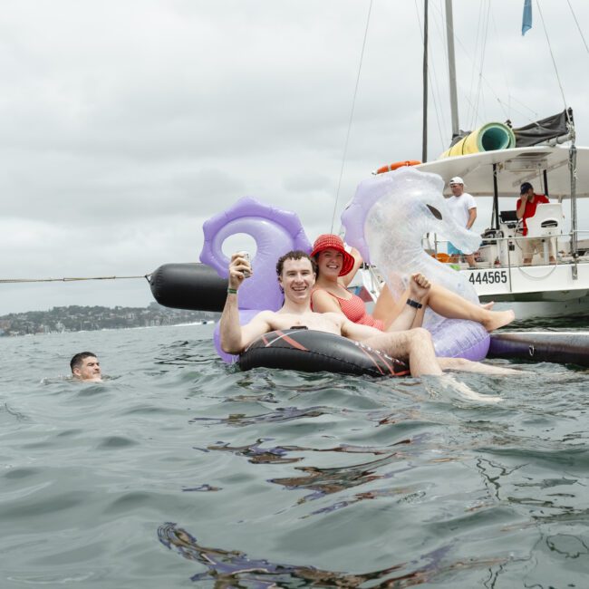 A group of people relax on inflatable rafts in the water near boats. One person is holding a drink while lounging on a unicorn-shaped float. The sky is cloudy, and others are seen socializing and enjoying the water.