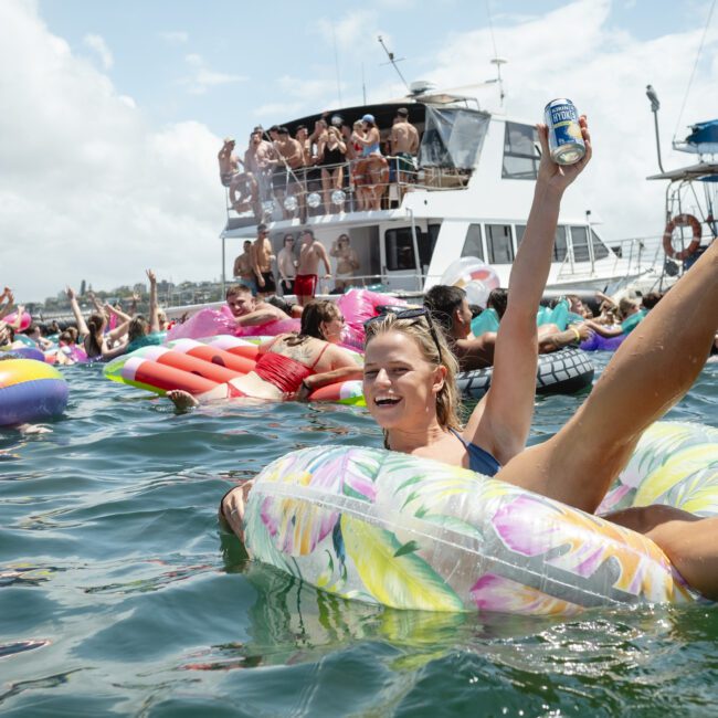 A woman in a swimsuit is joyfully floating on a colorful inflatable in the water, holding a can in one hand. A large group of people are on the water and aboard a boat, enjoying a sunny day.