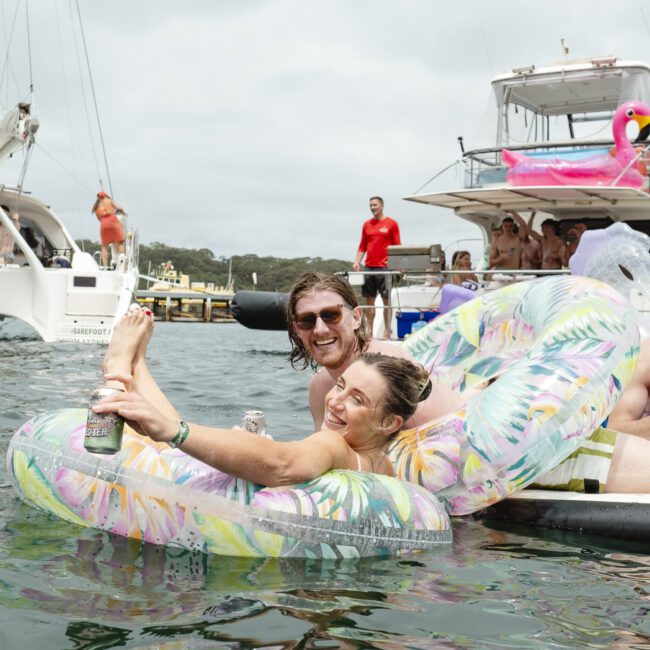 A man and woman relax on inflatable pool floats in the water, smiling and holding drinks. Behind them, a yacht with a large pink flamingo float and another boat are visible, along with partygoers. The scene is joyful and festive.