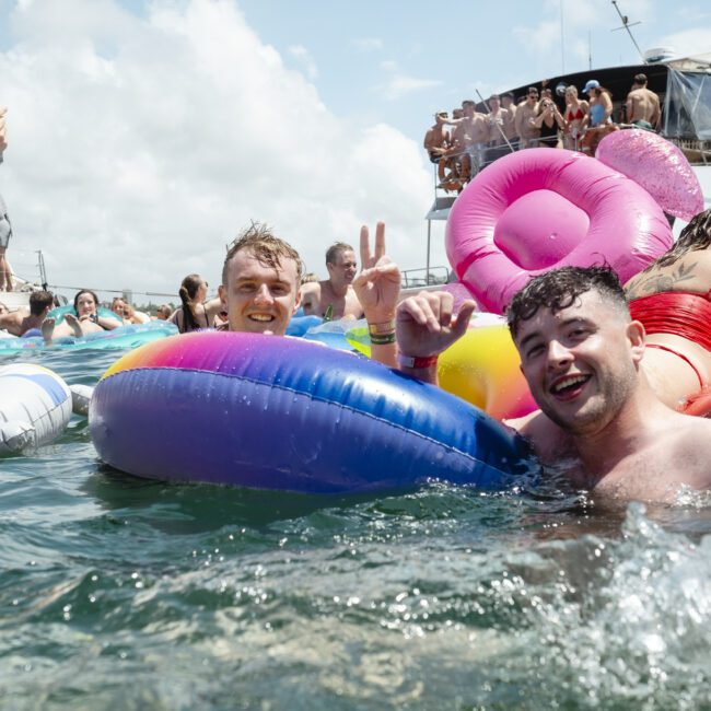 A group of people enjoys a sunny day on the water, lounging on colorful inflatable floats. Two men smile and gesture peace signs toward the camera. In the background, a large boat with more people is anchored nearby.