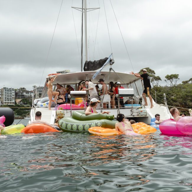 A group of people enjoy a day on the water, swimming and lounging on colorful inflatable floats near a yacht. The scene is set against a backdrop of greenery and buildings under a cloudy sky.