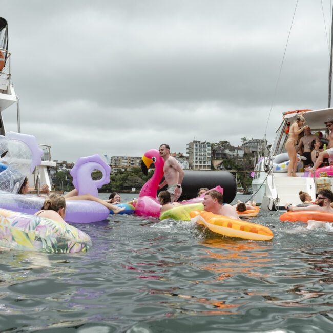 A group of people in a harbor are enjoying a day on the water. They are floating on colorful inflatables, including a flamingo and unicorn, near two boats. The sky is overcast, and buildings are visible in the background.