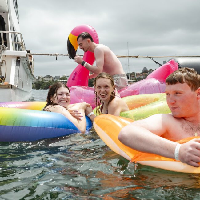 Four people enjoying themselves on colorful inflatable rafts in the water near a boat. They are smiling and appear relaxed, surrounded by large inflatables shaped like a flamingo and a unicorn.