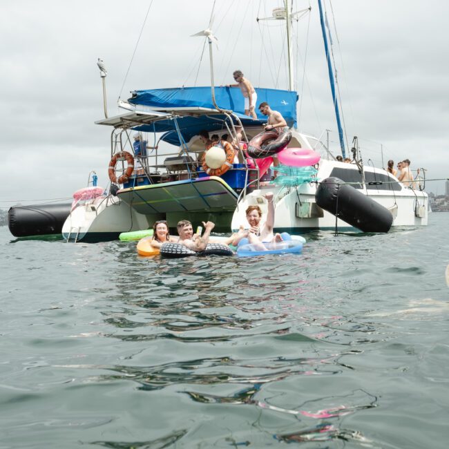 A group of people enjoying a day on the water near a sailboat. Some are lounging on inflatable rafts, while others are on the boat. The sky is overcast and the water is calm.