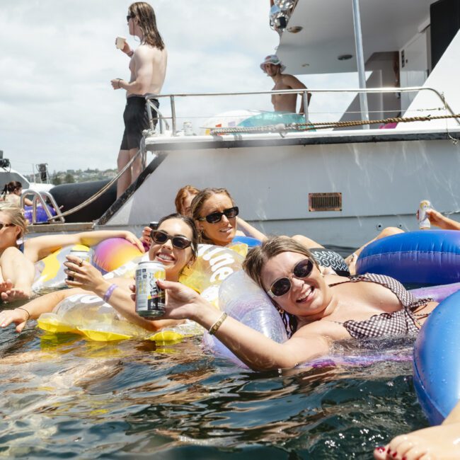 A group of people are lounging on colorful inflatable rings in the water near a boat. They are smiling and holding drinks, enjoying a sunny day. Other boats and more people can be seen in the background.