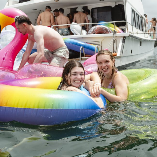 Two women smiling and relaxing on colorful inflatable floats in the water near a boat. One float is rainbow-striped, and the other resembles a flamingo. People can be seen on the boat in the background. The sky is overcast.