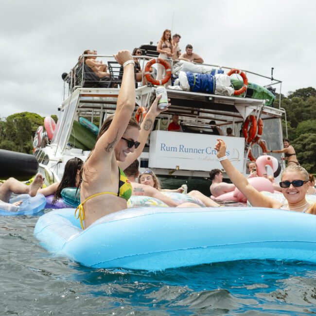 People are relaxing on inflatable floats in the water near a boat. The boat is crowded with more people having fun. Some are raising their arms in celebration. The scene is lively and festive, with a backdrop of trees and a cloudy sky.