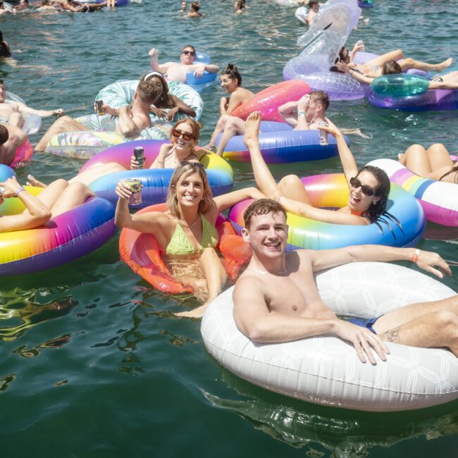 A group of people relaxing on colorful inflatable tubes in a lake, enjoying the sun. Some hold drinks, while others socialize and smile. The water is clear and the atmosphere is lively and fun.