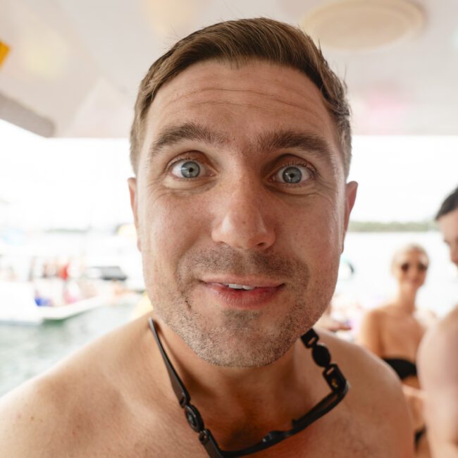 A man with short hair, wearing sunglasses around his neck, smiles into the camera on a boat. He is shirtless, and in the background, people relax near the water.