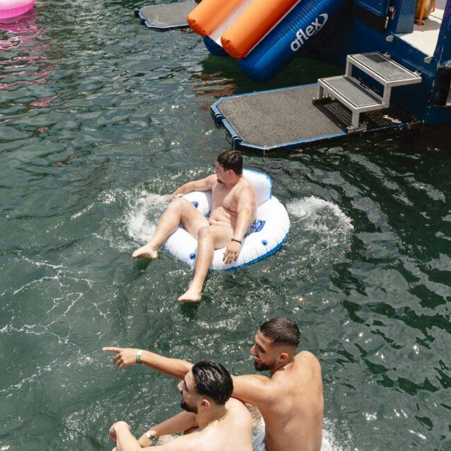 Two people sit on a dock, one pointing at something in the water. A person floats nearby in an inflatable ring. In the background, there is an inflatable slide leading into the water, with others gathered around a boat.