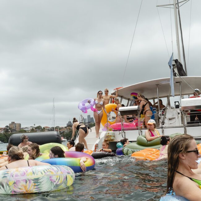 A large group of people enjoying a party on a boat and in the water, surrounded by colorful inflatable tubes and rafts. The sky is overcast, and the scene is lively with smiling attendees. The backdrop includes buildings and a marina.