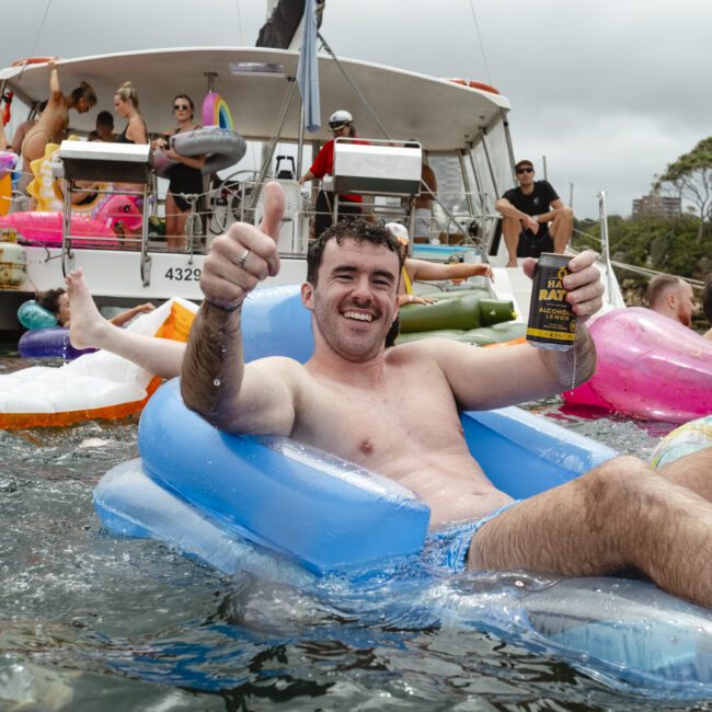 A man relaxes on an inflatable chair in the water, giving a thumbs-up and holding a drink. Behind him, people are enjoying a party on a boat and various floaties. The atmosphere is lively and festive, with a cloudy sky overhead.