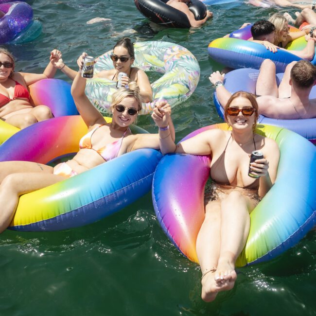 A group of people relax on colorful inflatable pool floats in the water, holding drinks and smiling. The sun is shining, and everyone appears to be enjoying a fun day.