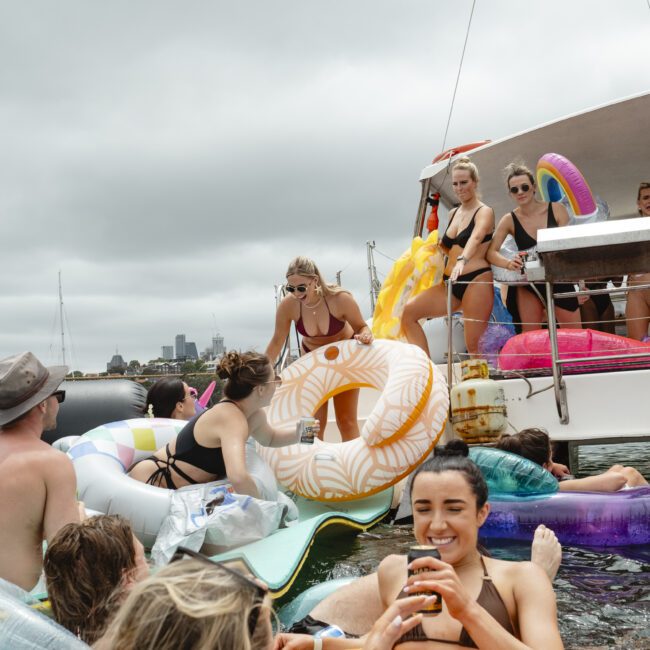 A group of people enjoying a sunny day on a boat and in the water. They are surrounded by colorful inflatables, including a donut and swan. Some are relaxing on floats while others are on the boat, with a city skyline in the background.