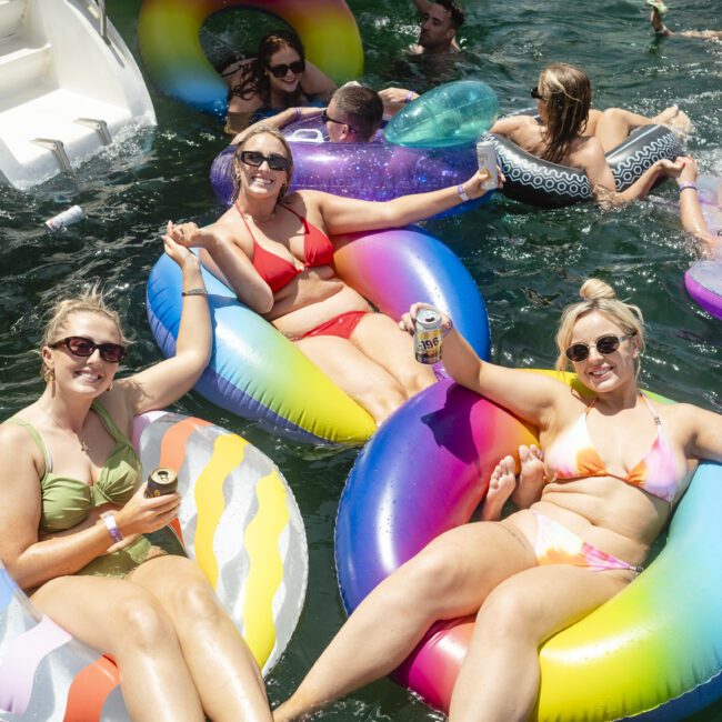 A group of people enjoying a sunny day in the water, lounging on colorful inflatable tubes. Three women in swimwear are smiling and holding drinks, with others swimming or relaxing nearby.