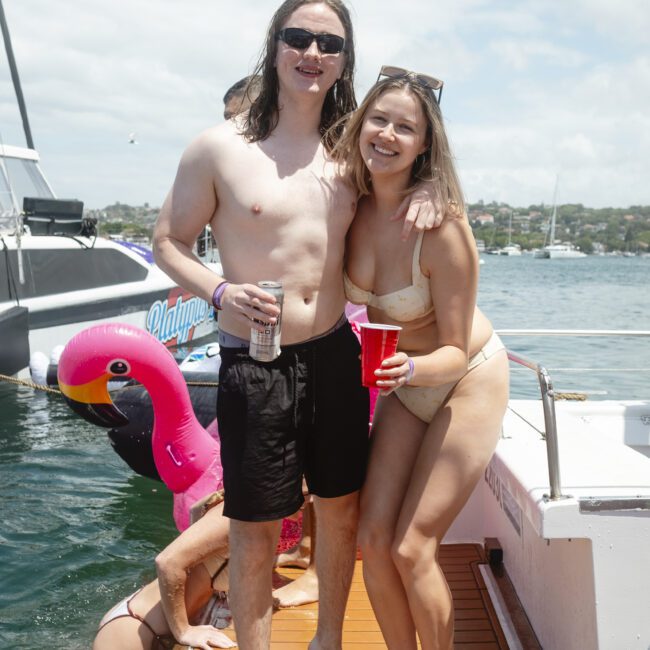 A smiling man and woman in swimsuits pose on a boat deck. The man holds a can and the woman holds a red cup. A pink flamingo float and water are visible in the background, with another person climbing onto the boat.