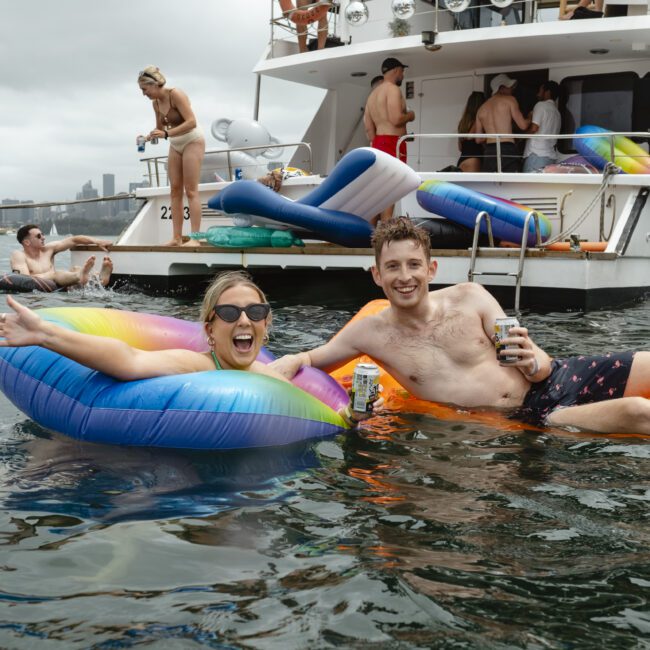 Two people relax on colorful inflatables in the water near a boat. They smile and hold drinks, surrounded by others enjoying the scene. The boat is equipped with more inflatables and people are gathered on the deck. The sky is overcast.