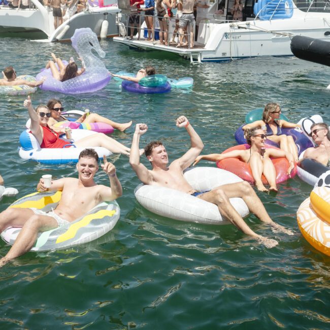 A group of people enjoying a sunny day on a river, lounging on various colorful inflatable tubes. Boats and more people are in the background. The atmosphere is lively and festive.