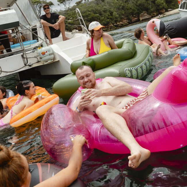 A group of people in swimsuits are lounging on colorful inflatable rafts in the water near a boat. One man is holding a drink on a pink flamingo-shaped raft. The scene is relaxed and joyful, with trees visible in the background.