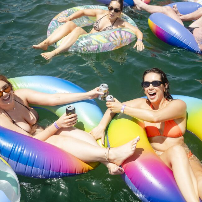 Two women in bikinis are floating on colorful inflatable rings in a lake. They're clinking cans and smiling. Other people are relaxing on inflatables in the background, enjoying a sunny day on the water.