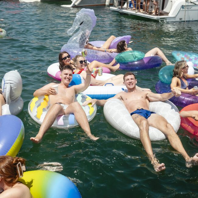 A group of people are floating on colorful inflatable rings and rafts in a sunny body of water. Some are relaxing, while others chat and enjoy drinks. A large boat is seen in the background. The atmosphere is lively and festive.