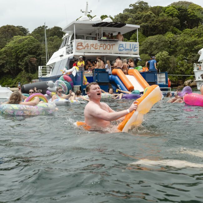 People enjoying a pool party on floaties and inflatable toys in the water near two boats named "Barefoot Blue." Lush greenery is visible in the background.