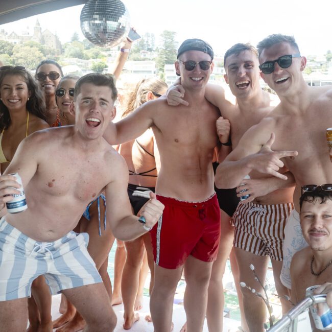 A group of people in swimwear enjoy a boat party, smiling and holding drinks. A disco ball hangs overhead, and trees are visible in the background. The atmosphere is lively and cheerful.