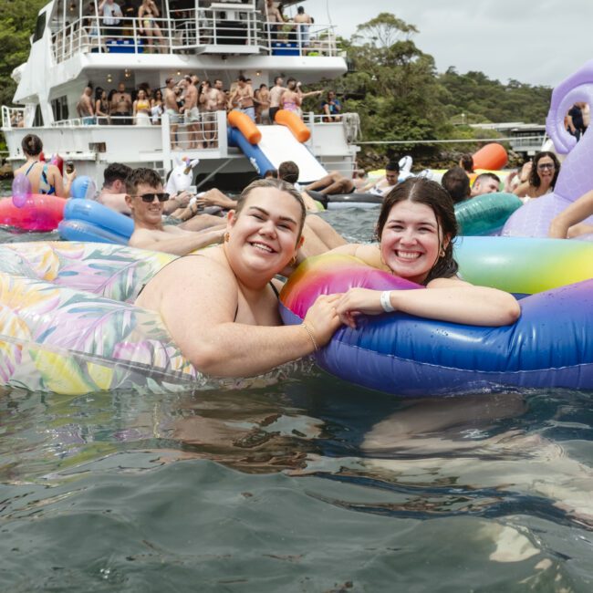 Two smiling people floating on colorful inflatable rings in the water. In the background, a boat with people on board is visible, along with trees on the shore. The atmosphere is lively and fun.