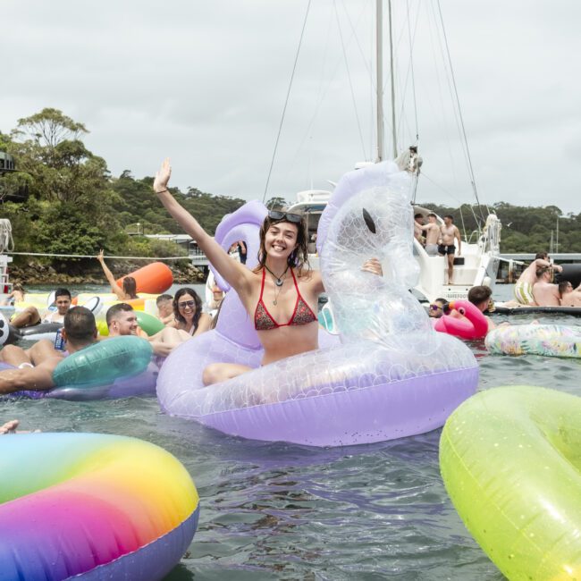 A person smiling and waving while sitting on a floating inflatable unicorn in a crowded water scene. Others are enjoying the water with various colorful inflatables. Boats and trees are visible in the background.