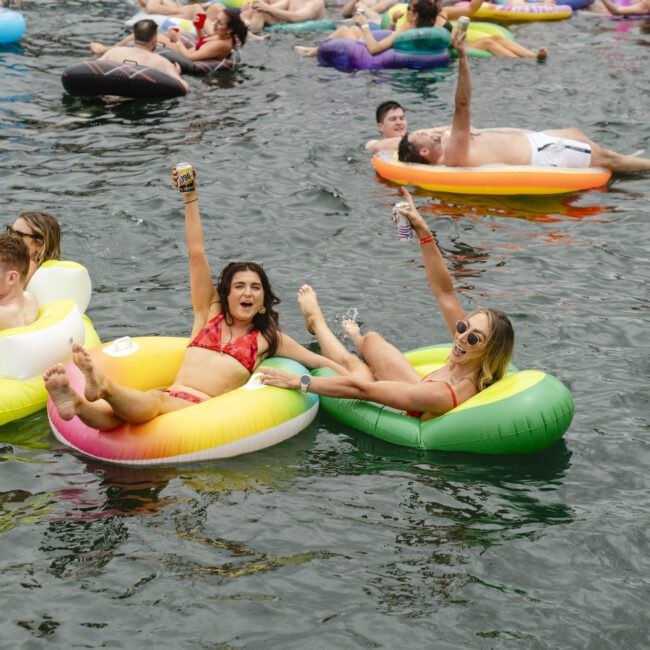 People enjoying a sunny day on a lake, relaxing on colorful inflatable tubes. They're holding up drinks and smiling, surrounded by others also lounging on the water. The atmosphere is lively and festive.