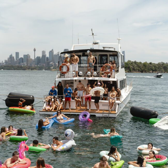 A group of people are enjoying a sunny day on a boat and in the water, surrounded by colorful inflatable floats shaped like watermelons and unicorns. The skyline is visible in the background.