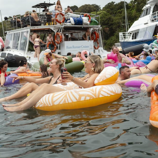 A lively scene on the water with people relaxing on colorful inflatables. Two boats in the background are filled with more people enjoying the day. The atmosphere is festive and social, with greenery visible in the distance.