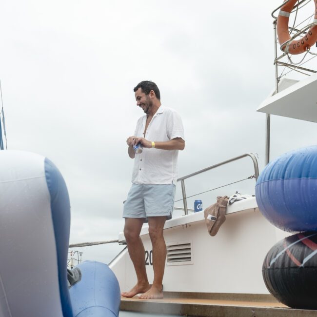 A man in a white shirt and shorts stands on the deck of a boat, next to large inflatable rings. The background features a cloudy sky and part of a sailboat.