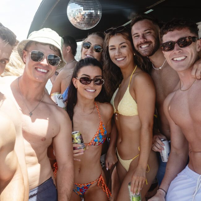 A group of smiling people in swimwear posing together on a boat, enjoying a sunny day. A disco ball hangs above them, and some hold canned drinks. The background shows clear skies.