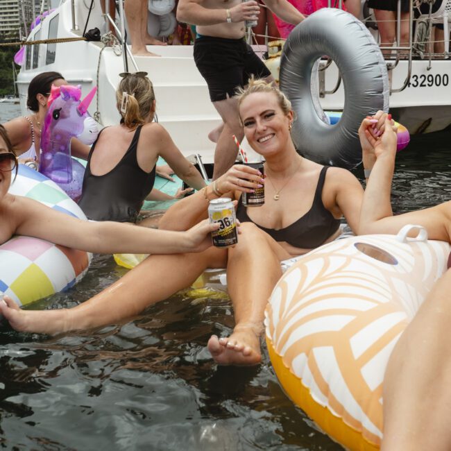 Three women in swimsuits enjoy a sunny day on a boat, sitting on inflatable floats in the water. They are smiling and holding drinks. Other people can be seen in the background on the boat and in the water.