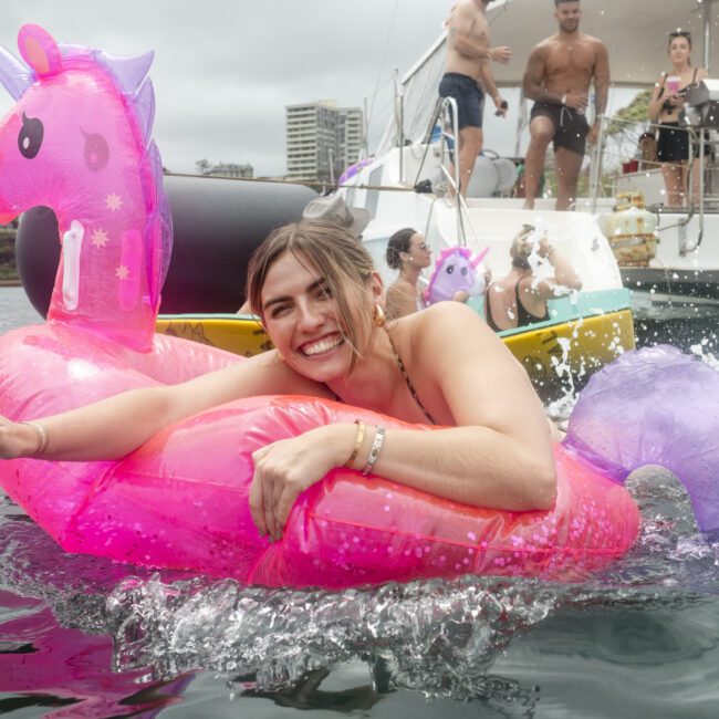 A person smiles while floating on a pink unicorn float in the water. A group of people is seen in the background on a boat, enjoying a casual day. Buildings are visible in the distance.