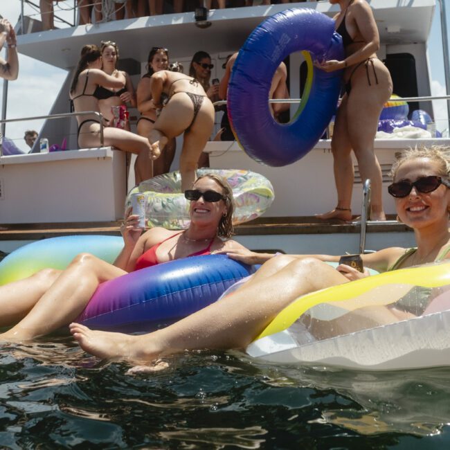 Two women relax on colorful inflatable pool floats in the water, holding drinks and smiling. A group of people in swimwear is on a boat in the background, with bright sunshine and blue skies above.