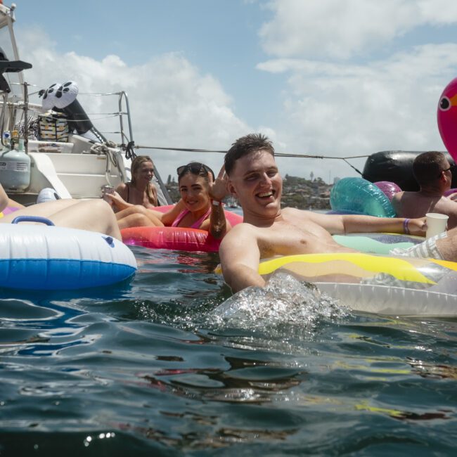 A group of people relax on inflatable floats, including a pink flamingo, in the water near a docked boat under a partly cloudy sky. They appear to be enjoying a sunny day, with one person holding a drink and smiling.