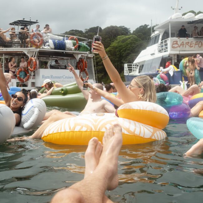 People relaxing on inflatable floats in a tropical water setting with a yacht nearby. A person is holding up a drink for a photo. Bright and sunny atmosphere with trees in the background.