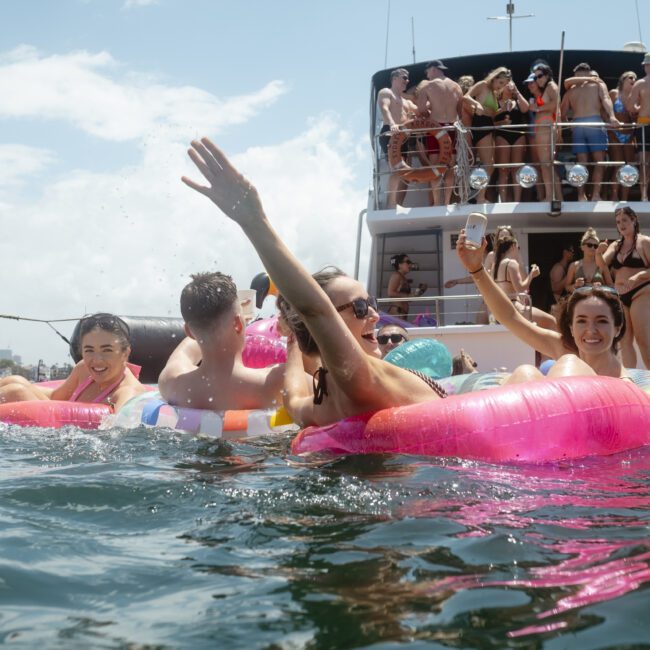 A group of people enjoy a sunny day on the water, floating with colorful inflatables. A boat nearby is filled with people socializing and taking photos. The scene is lively, with bright skies and city buildings in the background.